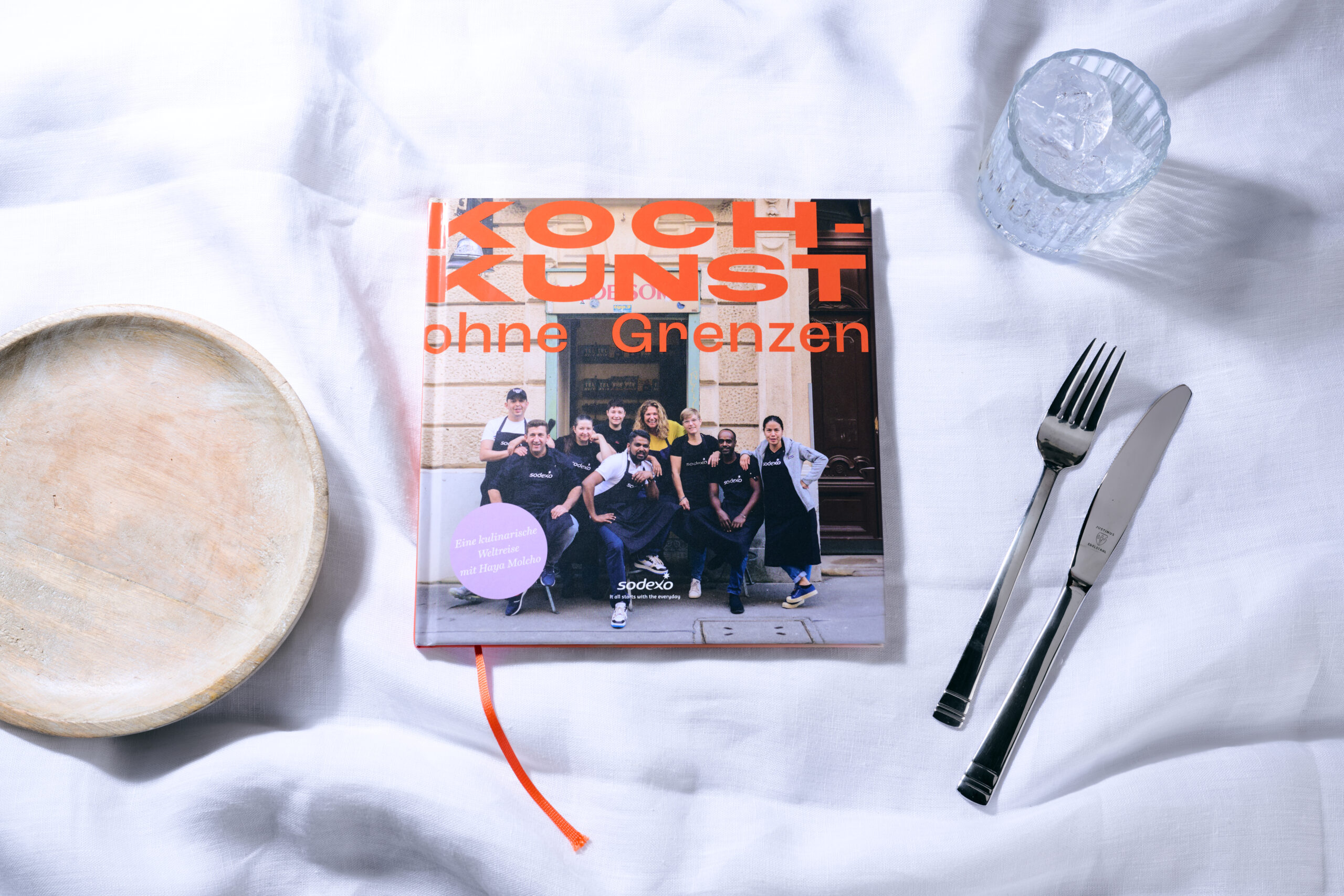 Kochbuch "Haya Molcho", Kochkunst ohne Grenzen, liegt auf einer Leinen-Tischdecke neben einem Teller, einem Glas und Besteck
